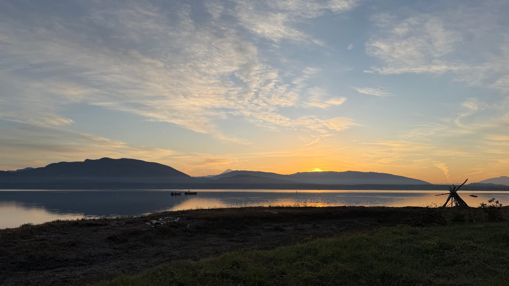 A beach in Saw, Washington. Sunset over some hills seen across a bay of water. A little stick structure is visible on the near shore.