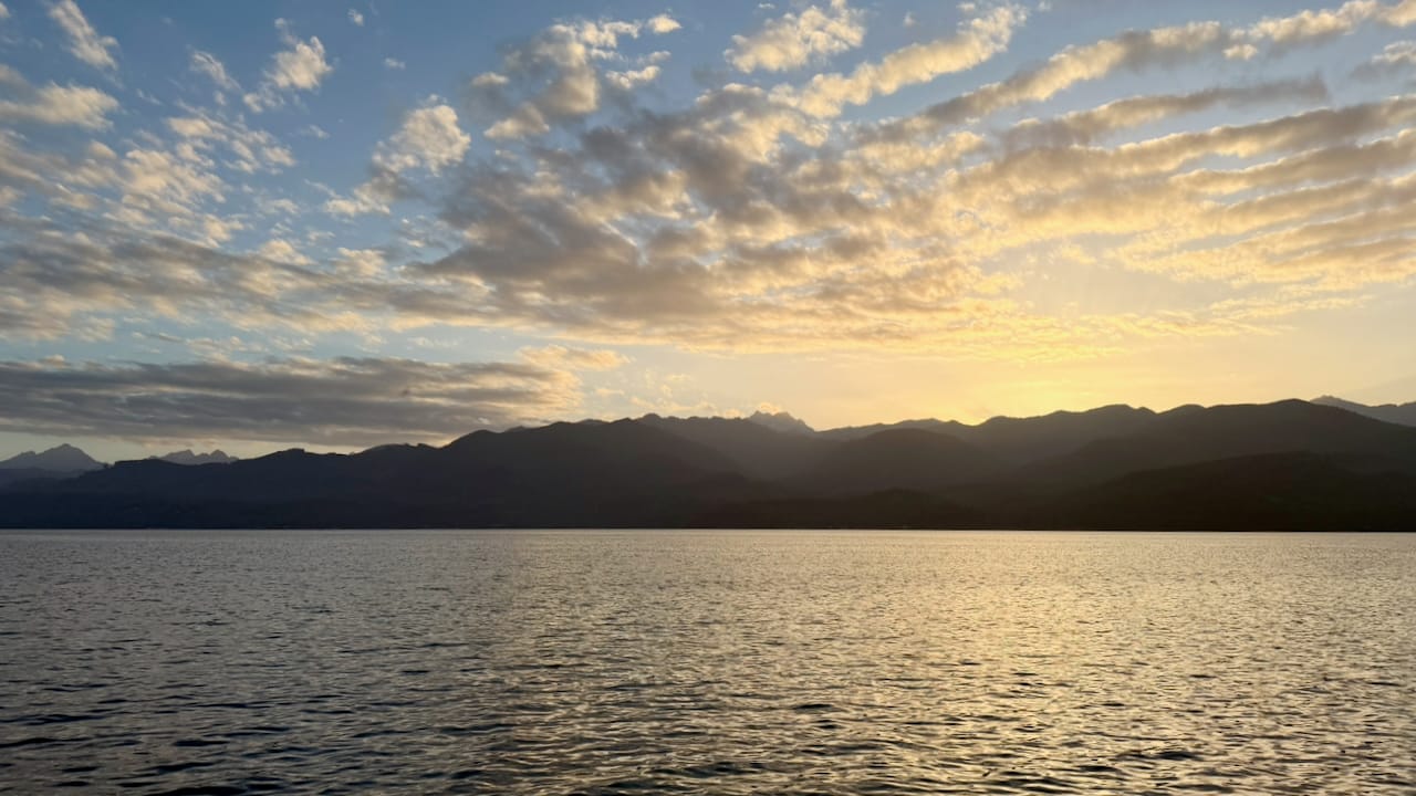 A sunset behind mountains across a body of water. There is a golden haze in the distance, reflected off the bottom of some clouds in the sky.