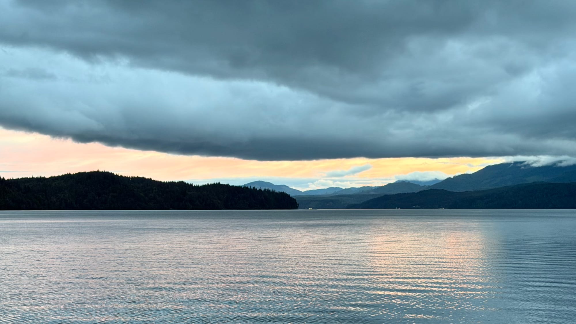Heavy clouds with the cliched silver linings moving across a body of water. Lighter skies can be seen off in the distance. 